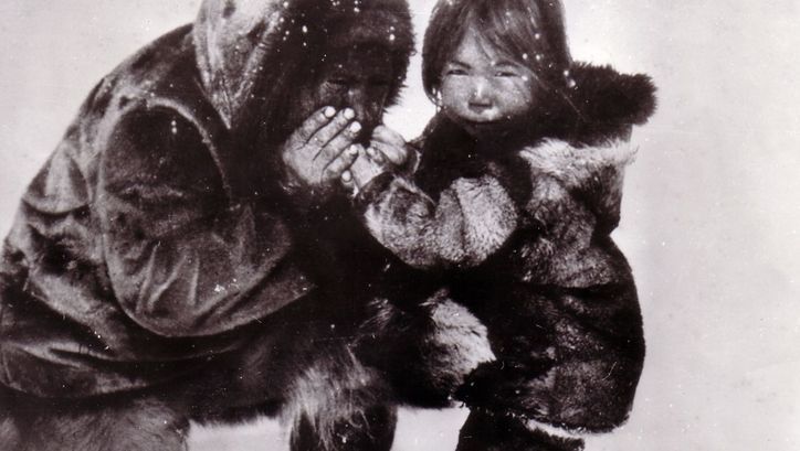 Film still from NANOOK OF THE NORTH. In a snowy landscape, a person warms the hands of a child.
