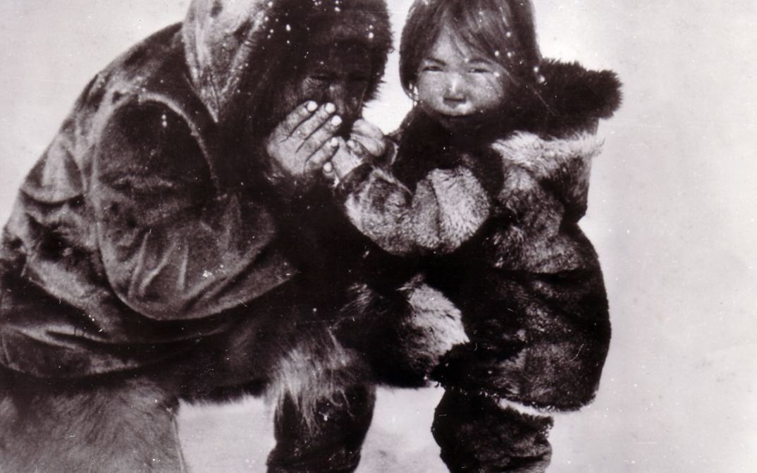 Film still from NANOOK OF THE NORTH. In a snowy landscape, a person warms the hands of a child.