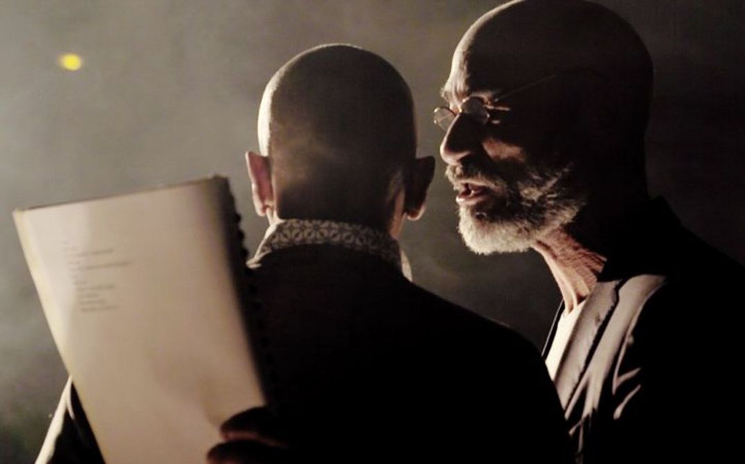 A close-up of two men. The first is standing with his back to the camera, the second can be seen in left side profile. It is dark and foggy. They are illuminated from the left.