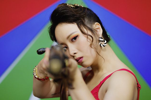Film still from THE TRIO HALL by Su Hui-yu. The image shows a close-up of a styled woman with a gun looking directly into the camera. The background consists of colorful stripes.