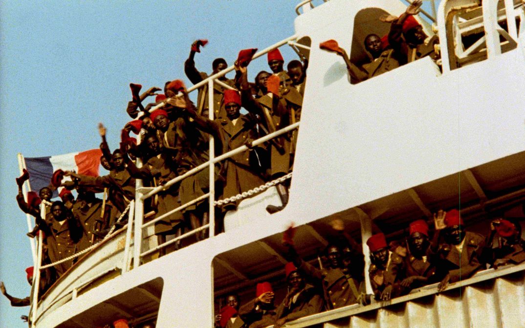 Filmstill from CAMP DE THIAROYE: People in long coats and red caps stand on a ship and wave. The French flag can be seen.