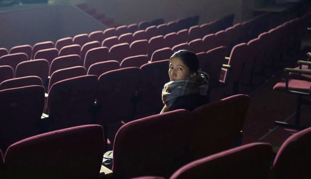 Filmstill aus „Akyn (Poet)“ von Darezhan Omirbayev. Eine Frau sitzt alleine in einem leeren Theatersaal. Sie dreht sich um und schaut in die Kamera. 