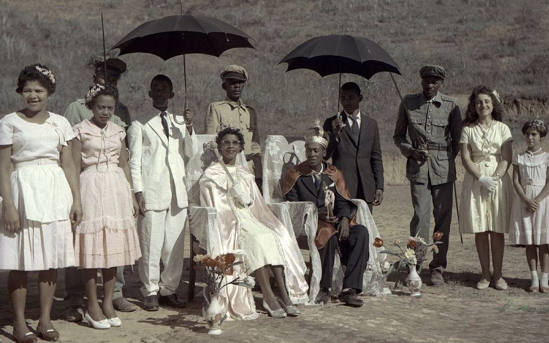 Film still from ALVORADA – AUFBRUCH IN BRASILIEN: A group of people are standing around two others, who are sitting on chairs, and holding two dark umbrellas.