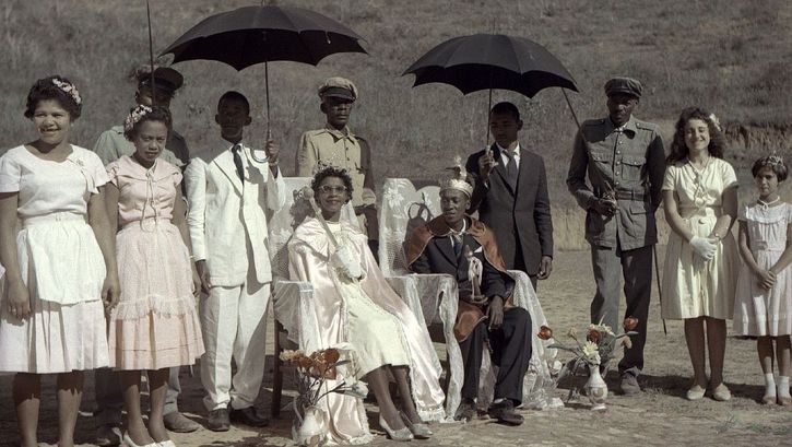 Film still from ALVORADA – AUFBRUCH IN BRASILIEN: A group of people are standing around two others, who are sitting on chairs, and holding two dark umbrellas.