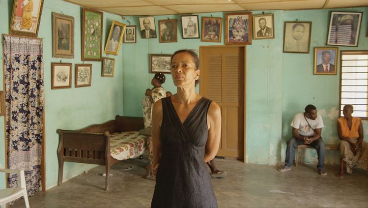 Filmstill aus BECOMING BLACK. Die Filmemacherin in einem Haus in Westafrika, sie schaut sich Fotos an der Wand an.