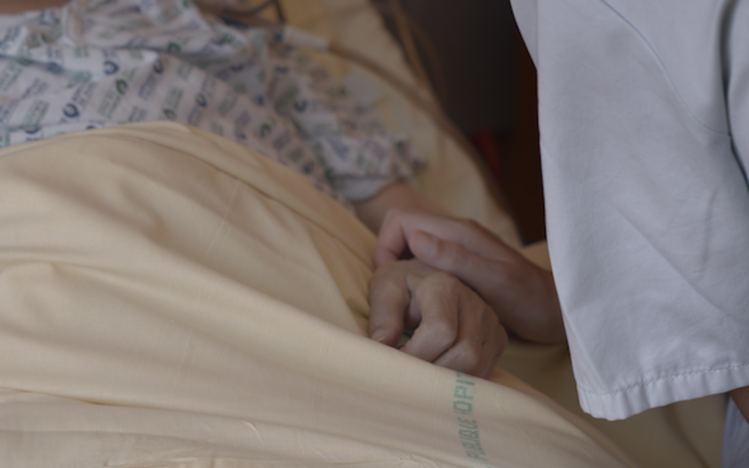Film still from NOTRE CORPS: One hand clasps another on a hospital bed.