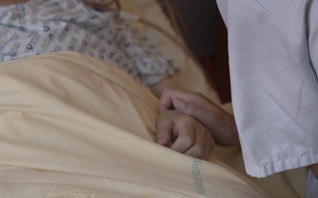 Film still from NOTRE CORPS: One hand clasps another on a hospital bed.