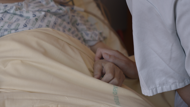 Film still from NOTRE CORPS: One hand clasps another on a hospital bed.
