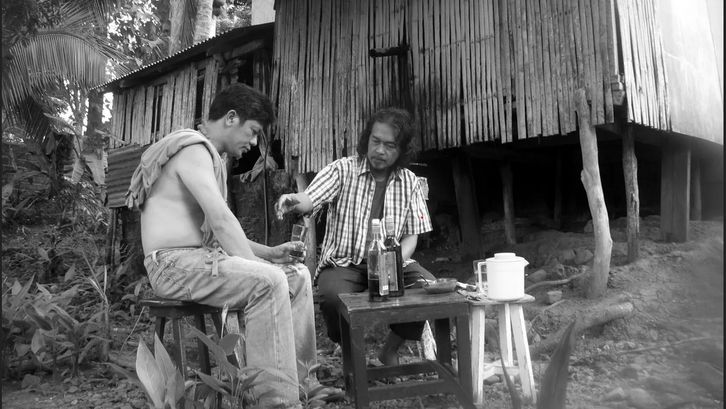 Filmstill aus BUTTERFLIES HAVE NO MEMORIES: Zwei Männer sitzen draußen vor einem Hütte an einem kleinen Tisch. Vor sich haben sie Getränke stehen.