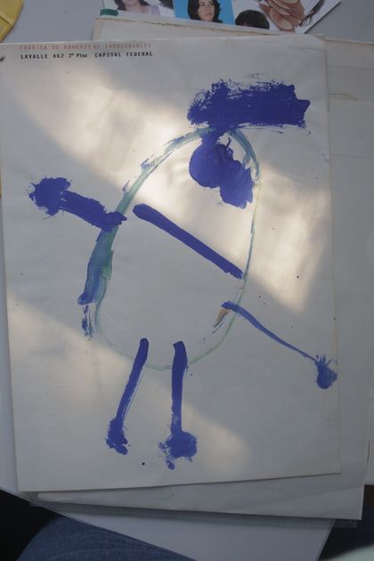 Eine Kinderzeichnung einer eiförmigen Figur mit blauer Farbe, mit Punkten als Augen, Linien als Arme, und einem Farbstreifen als Haare. Unter dem Papier sehen wir fragmentiert andere Dokumente, unter anderem Schulfotos.