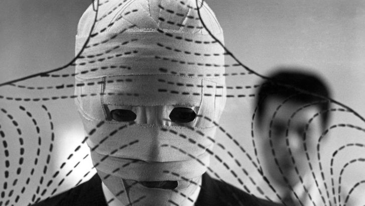 Film still from THE FACE OF ANOTHER: A man with a bandaged face stands in front of a transparent wall on which lines and strokes have been drawn.