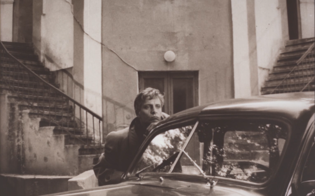 Film still from LJUDYNA K: A man leans against a car in front of steps.