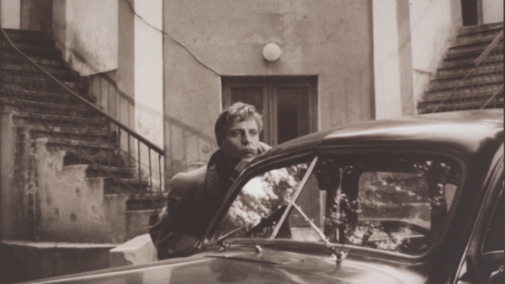 Filmstill aus LJUDYNA K: Vor Treppenstufen lehnt sich ein Mann an ein Auto.