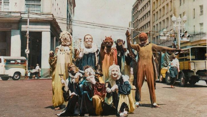 Filmstill aus PICTURES OF GHOSTS: Auf einer Straßenkreuzung posiert eine Gruppe von Menschen mit kunstvollen Masken und Kostümen.