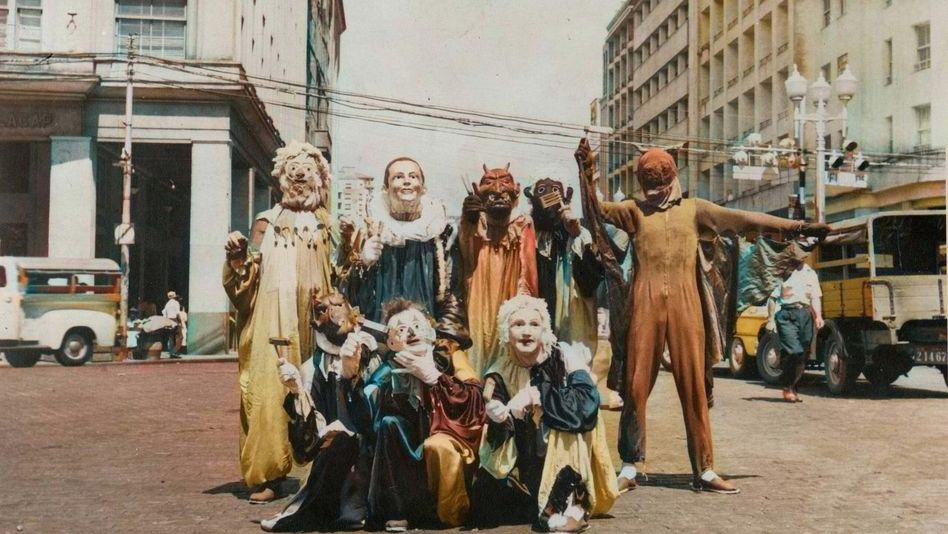 Film still from PICTURES OF GHOSTS: A group of people wearing elaborate masks and costumes pose at a crossroads.