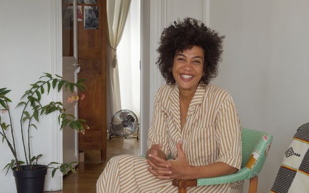 Eine Frau sitzt auf einem Stuhl in einem hellen Raum und spricht gestikulierend, im Hintergrund Pflanzen und eine offene Tür.