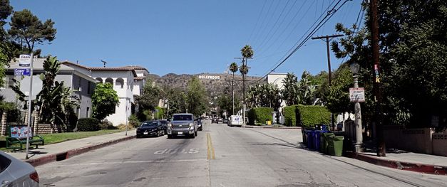 A two-lane road with houses and cars on the left and right. In the background is a hill on which the Hollywood sign can be seen. 