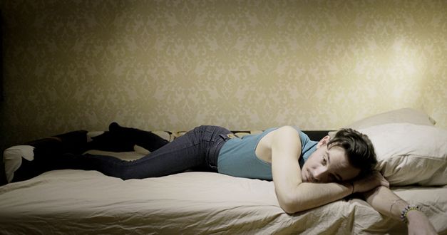 Filmstill aus „Llamadas desde Moscú“ von Luís Alejandro Yero. Ein Mann in Jeans und Top liegt auf dem Bauch in einem Bett und hat die Augen geöffnet.