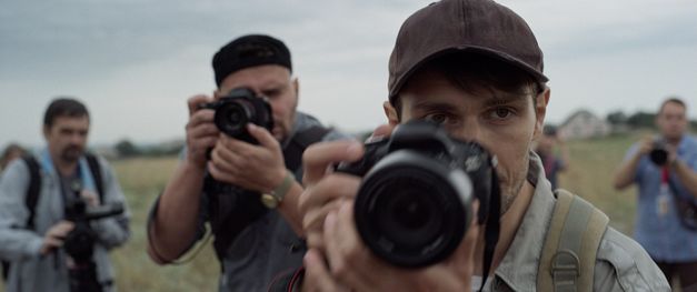 Film still from "Redaktsiya" by Roman Bondarchuk. It shows a close-up of people with photo cameras, all looking in the direction of the camera. Nature and a house can be seen in the background. 