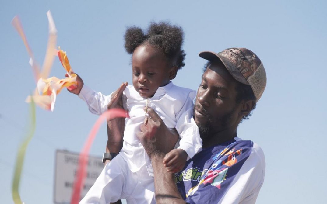 Ein Mann hält Kleinkind im Freien, bunte Bänder flattern im Wind.