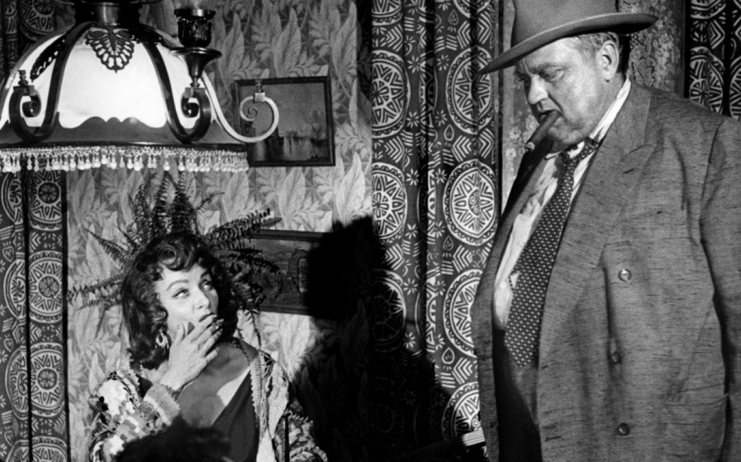 Filmstill from TOUCH OF EVIL: A man with a top hat, a cigar in his mouth and a suit stands next to a seated woman who is also smoking. In the background, patterned wallpaper, curtains and a steelyard.