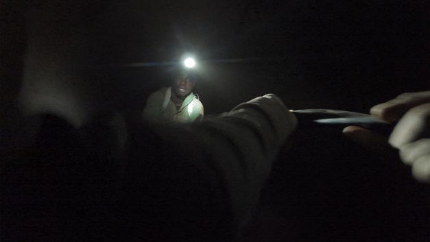 Still from the film "The Zama Zama Project" by Rosalind Morris. A man sits in the dark with a lit headlamp.