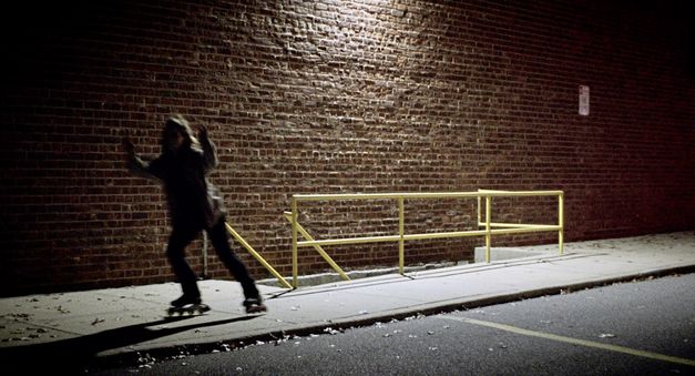 Still from the film "Happer’s Comet" by Tyler Taormina. A person on rollerblades rides past a basement entrance with a yellow railing at night.