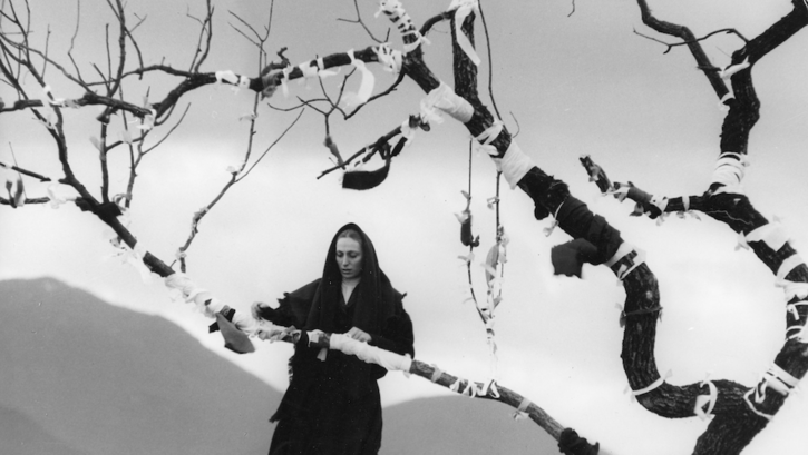 Film still from DAY IS LONGER THAN NIGHT: A woman dressed in black wraps ribbons around the branches of a tree.
