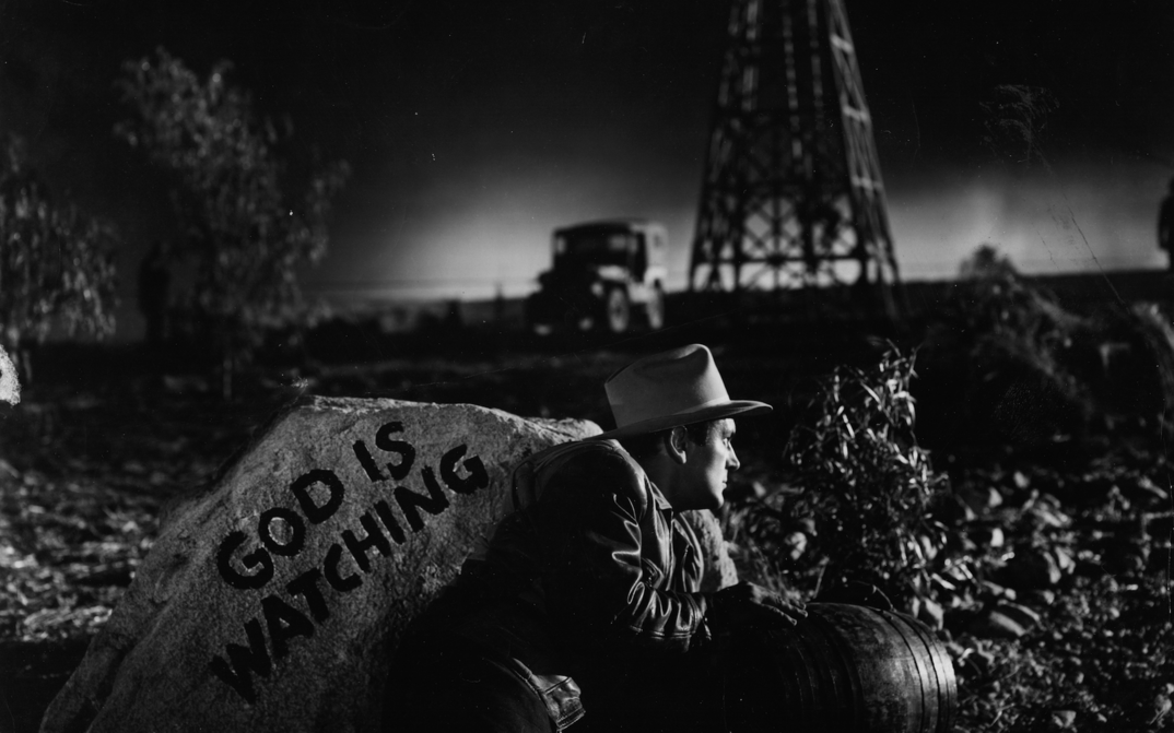 Film still from ESPALDAS MOJADAS: A man hides in the darkness behind a rock with the inscription "God is watching".