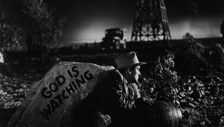 Film still from ESPALDAS MOJADAS: A man hides in the darkness behind a rock with the inscription "God is watching".