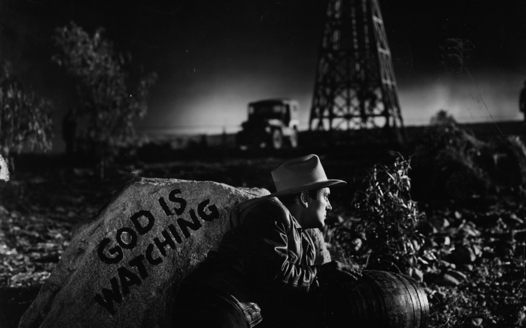 Film still from ESPALDAS MOJADAS: A man hides in the darkness behind a rock with the inscription "God is watching".
