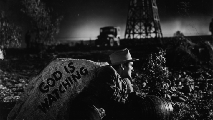 Filmstill aus ESPALDAS MOJADAS: Ein Mann versteckt sich in der Dunkelheit hinter einem Felsen mit der Aufschrift "God is watching".