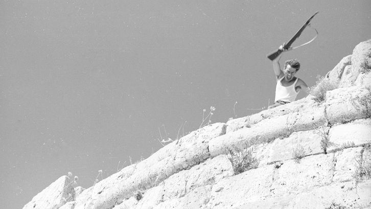 Film still from LEBENSZEICHEN: Above a rocky outcrop, a man is waving a rifle.