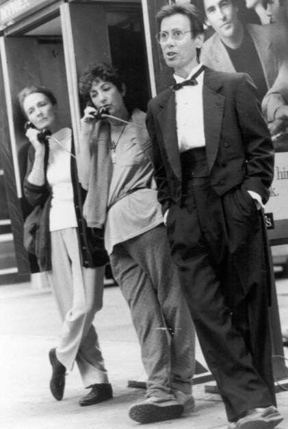 Set photo of MURDER and murder: director Yvonne Rainer stands in a suit next to her actresses, who are talking on the phone in phone booths.