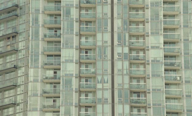 Filmstill aus „Mis Dos Voces“ von Lina Rodriguez. Frontale Aufnahme eines großen Wohnkomplexes. Die Fassade ist voller großer Fenster und Balkone, alle in leicht bläulichem Glas.