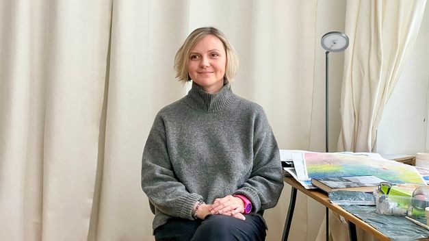 Eine blonde Frau sitzt in einem Raum neben einem Tisch mit Zeichnungen und Materialien, hinter ihr helle Vorhänge.