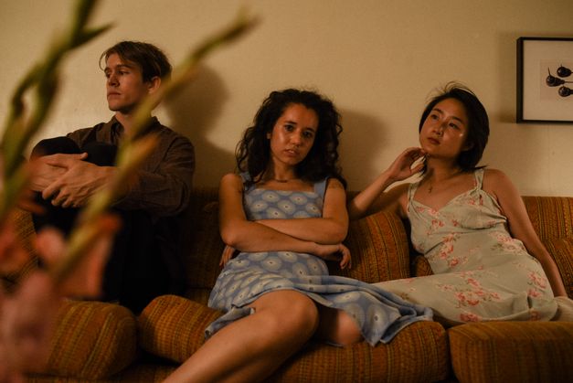 Filmstill aus ACTUAL PEOPLE. Drei junge Menschen sitzen auf einem Sofa und schauen in unterschiedliche Richtungen. Im Vordergrund ist unscharf eine Zimmerpflanze zu sehen.