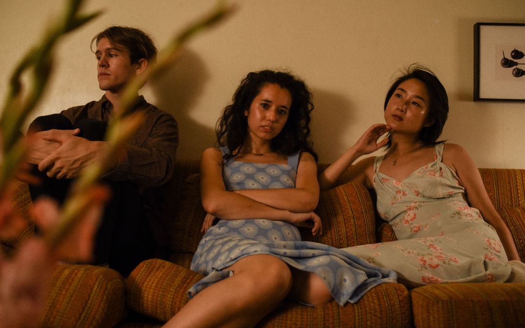 Film still from ACTUAL PEOPLE. Three young people sit on a sofa and look in different directions. In the foreground a plant can be seen out of focus.