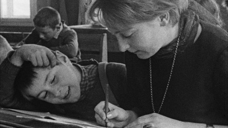 Film still from AUFSÄTZE: A teacher and a pupil are sitting in a school desk, she is writing something, he is watching.