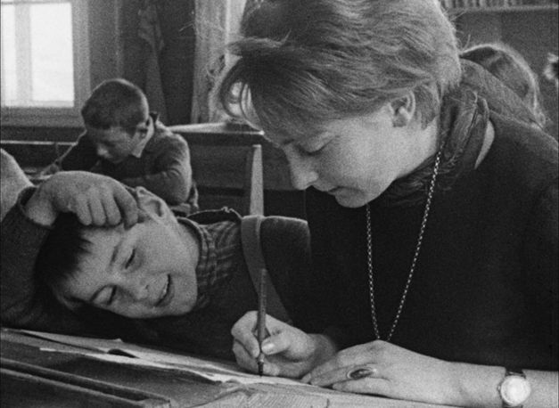 Film still from AUFSÄTZE: A teacher and a pupil are sitting in a school desk, she is writing something, he is watching.