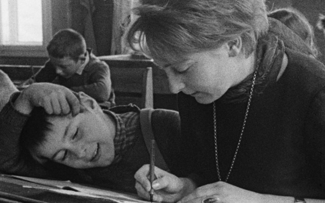 Film still from AUFSÄTZE: A teacher and a pupil are sitting in a school desk, she is writing something, he is watching.