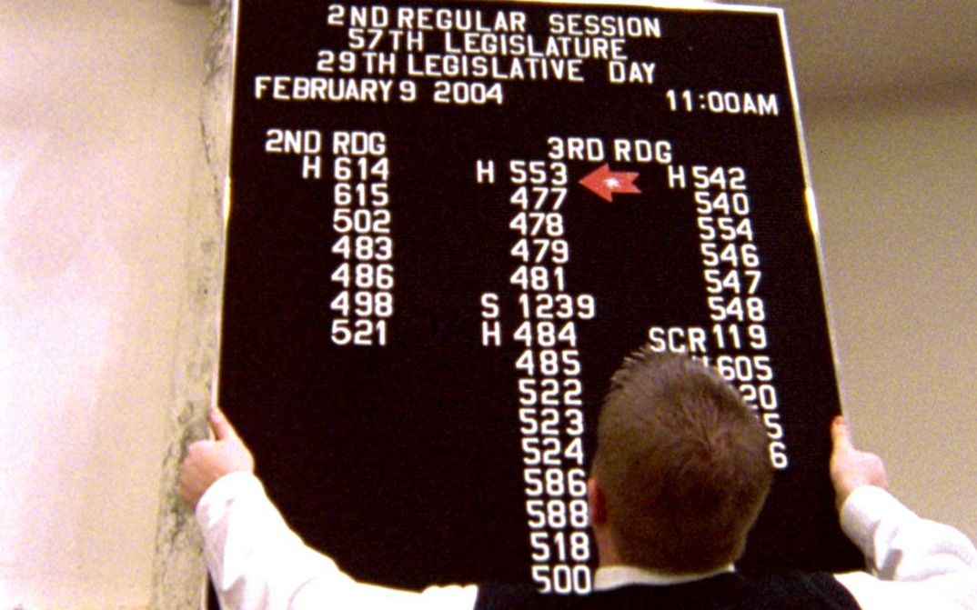 Film still from STATE LEGISLATURE. A man hangs an informational plaque at a legislative session in Boise, Idaho.