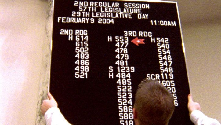 Filmstill aus STATE LEGISLATURE. Ein Mann hängt eine Informationstafel bei einer Parlamentstagung in Boise, Idaho auf.