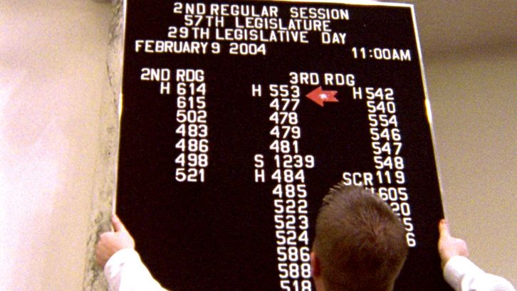 Film still from STATE LEGISLATURE. A man hangs an informational plaque at a legislative session in Boise, Idaho.