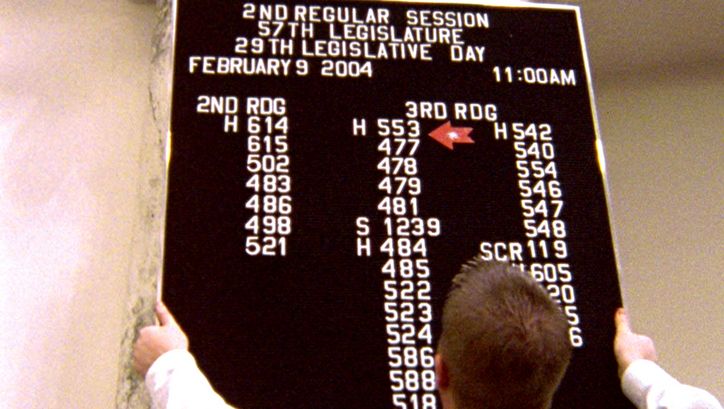 Filmstill aus STATE LEGISLATURE. Ein Mann hängt eine Informationstafel bei einer Parlamentstagung in Boise, Idaho auf.
