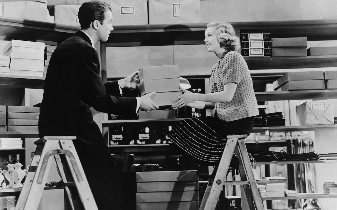 Film still from THE SHOP AROUND THE CORNER: A man and a woman are sitting opposite each other on two ladders. They are in a room full of shelves and boxes and are passing some boxes to each other.