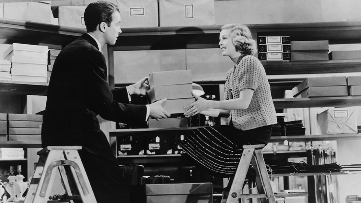 Film still from THE SHOP AROUND THE CORNER: A man and a woman are sitting opposite each other on two ladders. They are in a room full of shelves and boxes and are passing some boxes to each other.