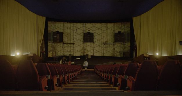 Filmstill aus „Scala“ von Ananta Thitanat. Eine Aufnahme aus dem hinteren Teil eines Kinos nach vorne. Die Leinwand ist schon abgebaut, wir sehen nur das Gerüst und die Lautsprecher. Ein Man läuft die zentrale Treppe hinunter. 