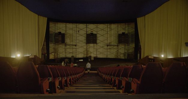 Still from the film "Scala" by Ananta Thitanat. In a shot from the back of a cinema looking forward, we see a man walking down the centre row. Where the screen should be are only beams and speakers. 