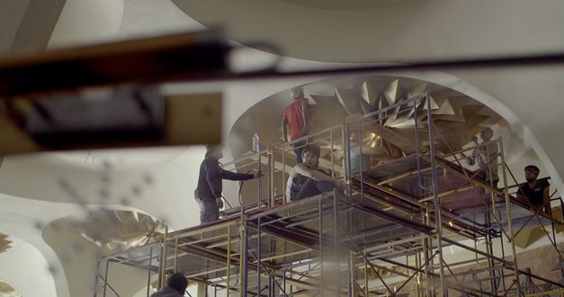 Still from the film "Scala" by Ananta Thitanat. We see a shot from below as a group of men on scaffolding work to remove light fixtures from the ceiling.